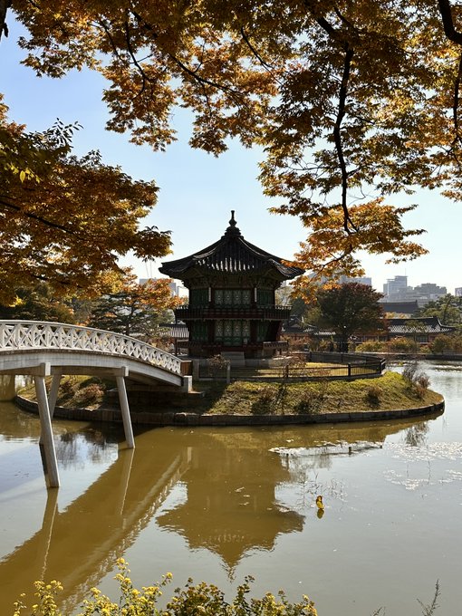 A traditional Korean pavilion sits on a small island surrounded by a tranquil pond, framed by a white arched bridge and golden autumn foliage, with a distant cityscape blending old and new architecture. The serene, historic setting evokes Korea's rich cultural heritage, setting the perfect tone for a post about attending Korea Blockchain Week 2025.