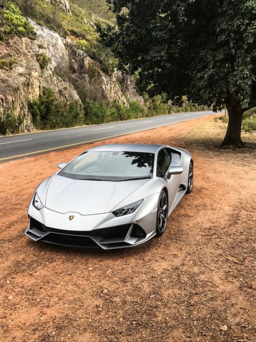 A silver Lamborghini Huracán sports car parked on a dirt area next to a road, with rocky hills and trees in the background.