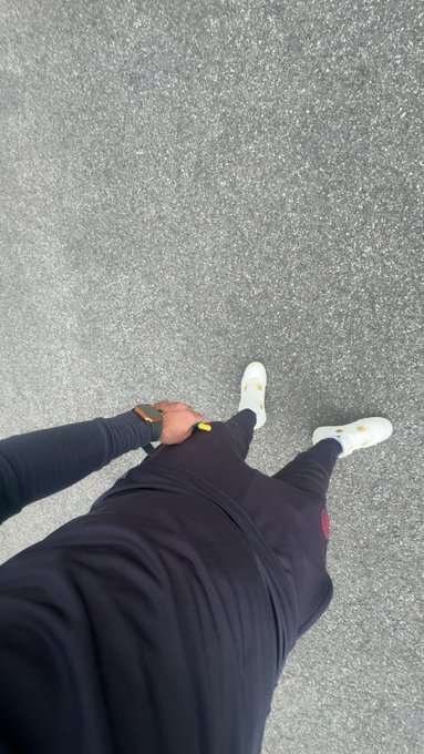 A first-person view looking down at the photographer's outfit—black joggers, a black long-sleeve top, and white sneakers with yellow accents, paired with a gold watch—standing on asphalt pavement, showcasing the casual streetwear "fit" being asked about in the caption.