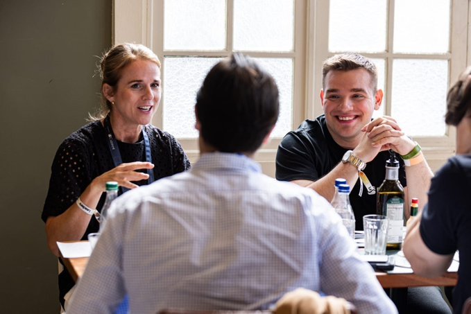 A group of colleagues engaged in a lively conversation around a table near a window, with drinks and a bottle visible.