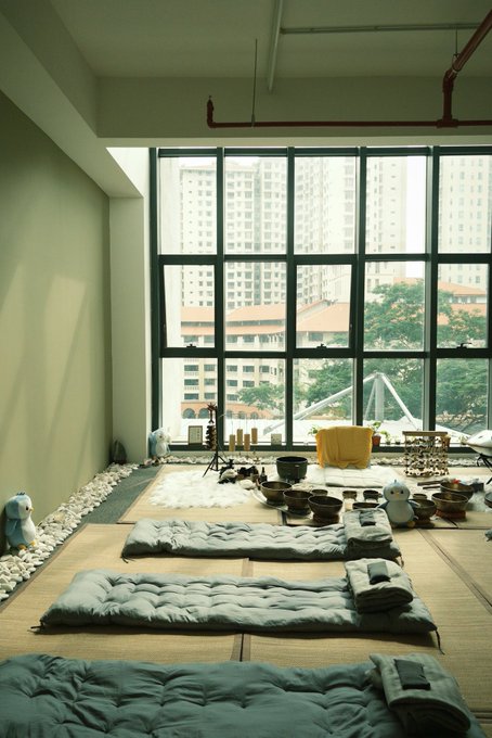 A serene wellness room with green sleeping mats and meditation cushions arranged on tatami flooring, surrounded by white stones and plush toys, with large windows overlooking residential buildings.