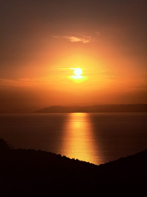 A vibrant orange sunset over a calm body of water, with the sun low on the horizon and distant hills silhouetted in dark tones.