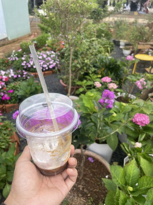 Hand holding a clear plastic cup with an iced beverage and straw in front of a garden filled with potted plants and pink and purple flowers.