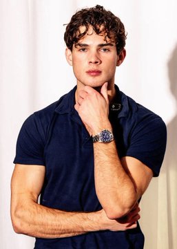 A young man with styled curly brown hair poses in a navy blue polo shirt, resting his hand near his chin to display a prominent wristwatch, exuding a confident, looksmaxing aesthetic that complements the satirical post about Clavicular's claims of plague immunity.