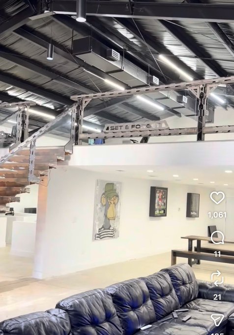 A spacious, modern loft-style studio features an industrial ceiling with exposed beams and ductwork, alongside a wooden staircase, a large plush black leather sofa, and artwork on white walls—including a Bored Ape Yacht Club piece. The post highlights the discovery of this Bored Ape artwork in the studio, celebrating the connection between the studio owner and the BAYC community.