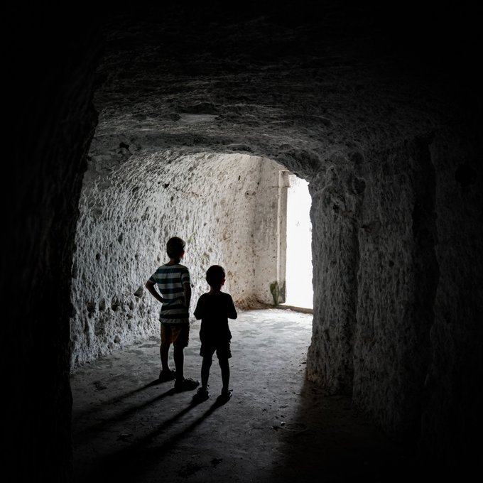 The silhouettes of two children stand in a dimly lit stone tunnel or cavern, looking toward a bright, sunlit opening at the end.