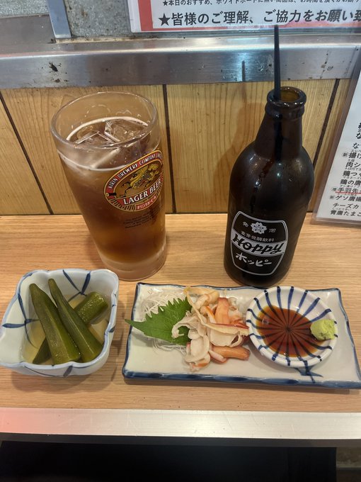A Japanese restaurant table setting featuring a glass of Kirin Lager Beer, a bottle of Hoppy (a Japanese beer-like beverage), pickled cucumbers, and a sashimi appetizer with shrimp and soy sauce.