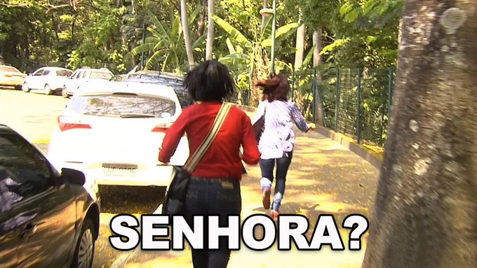 The focus of the image is two people walking down a path near trees, with cars parked nearby. They are heading towards a car that has a sign attached to its back that reads "senhora?". The backdrop features lush green trees and an outdoor setting, while the presence of various vehicles adds context to the scene. In the foreground on the image, there is text that reads "Minha cabeça vendo o ETH a $1500", possibly indicating that this photo may be associated with a marketing campaign or social media post about a recent event involving the two women and a car parked nearby.