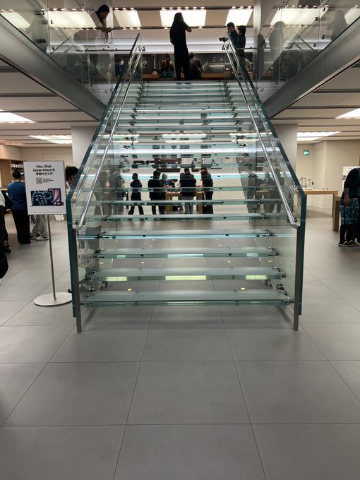 A glass staircase in what appears to be an Apple Store with people gathered on the upper level. The post's comment about "pants visible" seems to joke about how the transparent steps create an unintentionally revealing view of customers' legs from below.