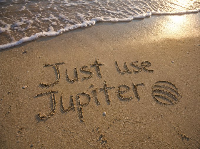 The phrase 'Just use Jupiter' written in the sand on a beach, accompanied by a drawing of Jupiter's striped appearance, with ocean waves washing ashore.