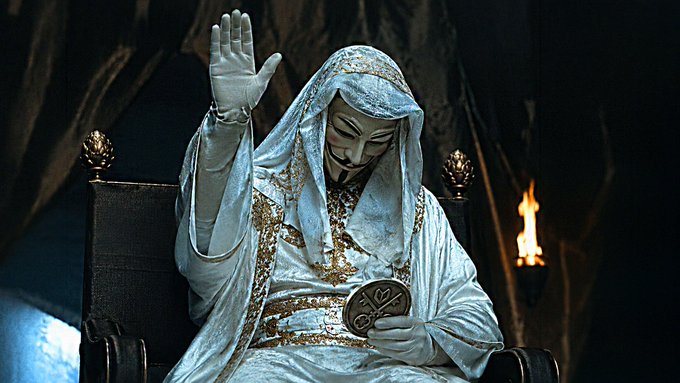 A figure dressed in ornate white and gold robes wearing a stylized mask raises one gloved hand while seated on a throne-like chair with flames visible in the background.