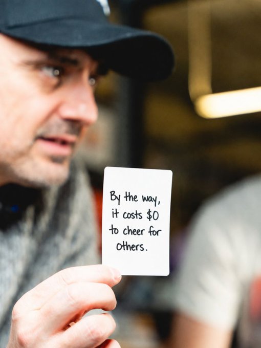 A man wearing a black cap holds up a small white card that reads, 'By the way, it costs $0 to cheer for others.'