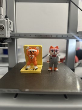 Two small figurines—an orange cat in a yellow photo frame labeled "Bricks" and a fox in a gray suit—sit on a 3D printer bed, likely representing a community giveaway theme from a "GM" (Good Morning) post. The "Bricks" branding suggests this is a promo for a crypto or NFT project, with the post encouraging engagement for a chance to win.