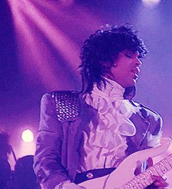 The image features a man playing an electric guitar, wearing a purple suit with a white shirt and ruffled collar. The man appears to be performing at a concert or event, as there are other people present in the background who might also be part of the performance.