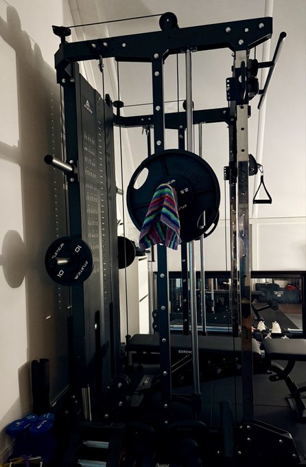 A cozy home gym setup featuring a black cable machine with weight plates, one of which is labeled "10" and another "45 lbs," alongside a colorful striped towel draped over the bar. The text "COREFX" is visible on the equipment, fitting perfectly with the "Good morning Home gym time" vibe of an early workout session.