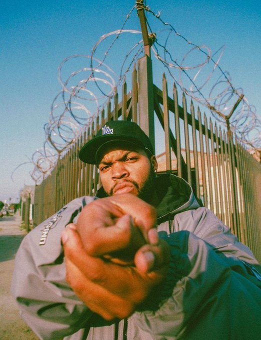 A photograph of a man wearing a '1of2' cap and gray jacket pointing directly at the camera, standing in front of a metal fence topped with coiled razor wire.