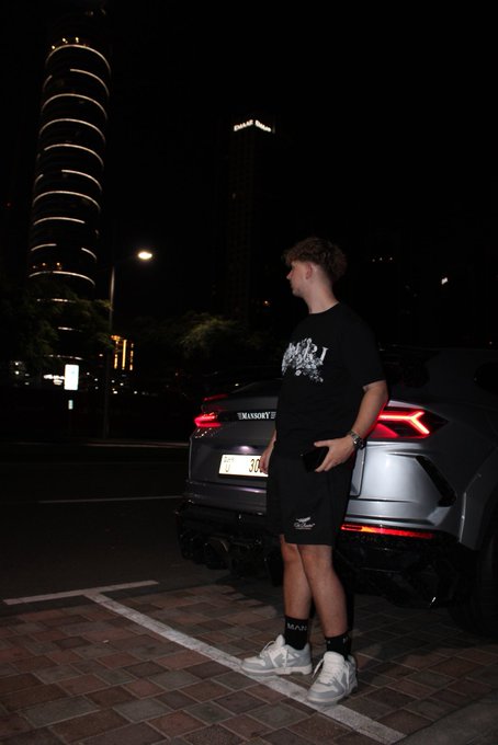 A young man in a black Karl graphic t-shirt and shorts stands next to a grey Lamborghini Urus at night with illuminated skyscrapers in the background.