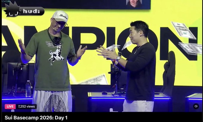 A YouTube livestream screenshot from 'Sui Basecamp 2026: Day 1' showing two men on stage with 'CHAMPION' text in the background, with money being thrown into the air.