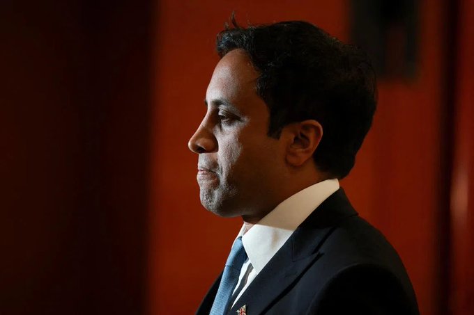 A profile portrait of a man in a dark suit and blue tie, photographed in side profile against a deep red background.