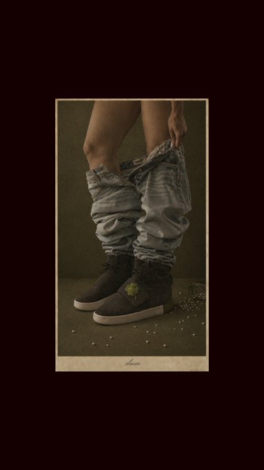 A stylized fashion photograph shows a person wearing loose acid-wash denim jeans and dark high-top sneakers with a green clover detail, with scattered debris on the floor.