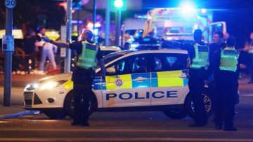 Two police officers in high-visibility vests stand beside a marked police car with its lights flashing at night, managing a scene while bystanders gather in the background. The visible "POLICE" text and emergency response presence connect to the post about the kidnapping and violent robbery of two French crypto millionaires in London, suggesting law enforcement involvement in the aftermath of the crime.