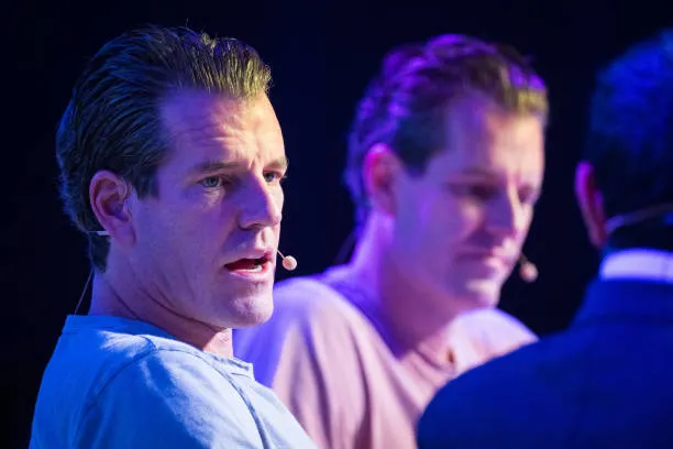 Two men wearing headset microphones are shown on a stage with blue lighting, appearing to be participating in a panel discussion or live show performance.