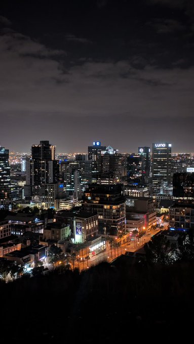 The image shows a city skyline at night with tall buildings illuminated by streetlights. The sky above the city appears dark, creating an interesting contrast between the bright lights of the buildings and the darkness below. The photo captures the essence of urban life and highlights the beauty of the cityscape under the cover of night.