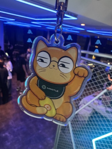 An acrylic keychain featuring an orange tabby cat character wearing a green bib that says 'MARSTLE' is being held up in what appears to be a neon-lit event space.