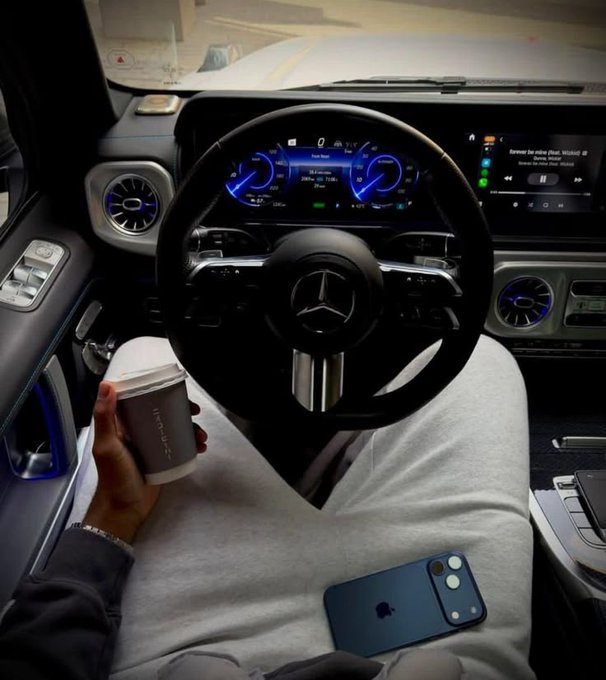 The image shows the interior of a Mercedes-Benz vehicle, with a person in the driver's seat holding a paper coffee cup (likely Starbucks) and an iPhone resting on their lap, with the dashboard display showing Bluetooth audio connectivity. The image relates to the post's casual, lifestyle-oriented vibe, suggesting a "breakfast on the go" morning routine in a luxury car.