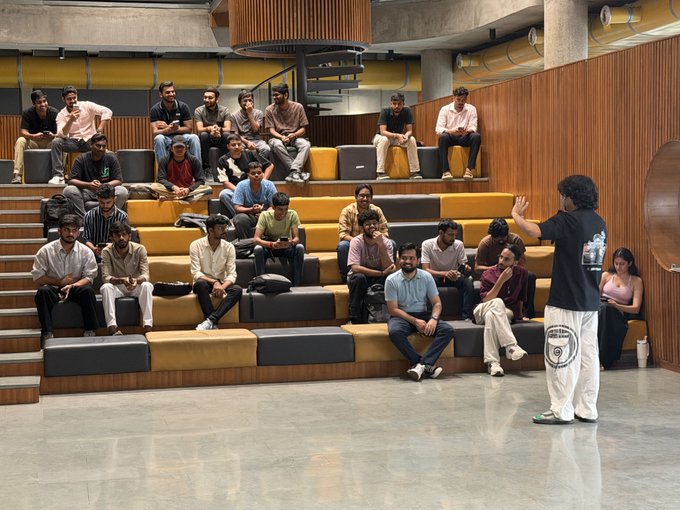 A group of people sits on tiered seating in a modern auditorium, listening to a presenter standing on the floor in front of them.