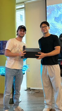 Two men smile while holding a box together at what appears to be a tech event, with the partial text "R" and "pools" visible on a presentation screen behind them, aligning with the post celebrating 200+ attendees at a Seoul gathering focused on the verUSD project.