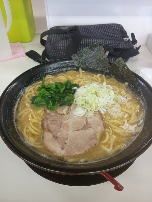 A bowl of ramen with chashu pork, green onions, spinach, and nori seaweed, with a bag and yellow container in the background.