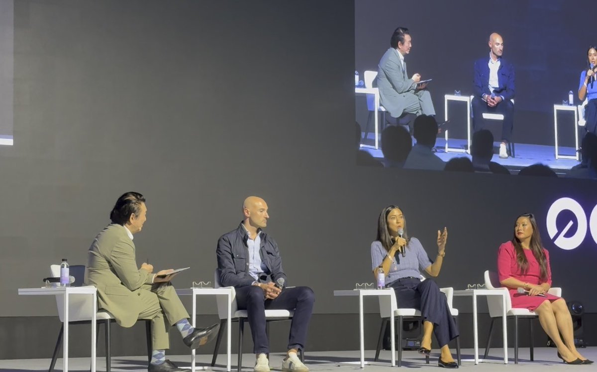 A panel discussion on stage with four people seated in chairs, shown both live and on a large screen in the background.