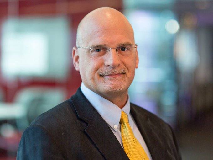 A professional headshot of a bald man with a goatee, wearing glasses, a dark suit jacket, a light blue shirt, and a yellow patterned tie, smiling at the camera.
