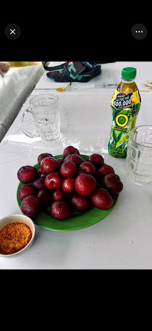 A plate of red plums or similar fruit on a green leaf, accompanied by a bottle of 'Number One' green tea, glasses of water, and a small dish of chili salt on a white table.