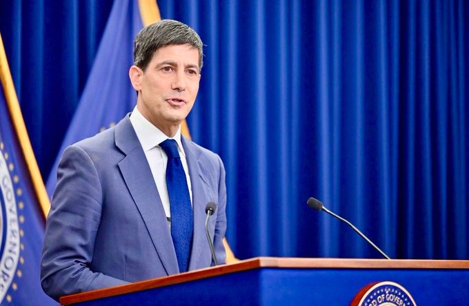 A man in a blue suit and tie stands at a podium with microphones, speaking in front of blue curtains and flags, with a partial 'Office' seal visible on the podium.