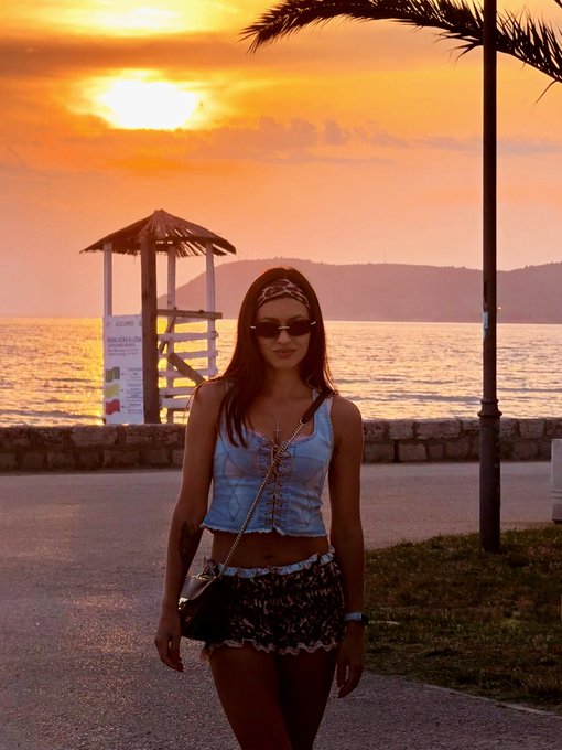 The image features a woman walking down a street at sunset, wearing sunglasses and carrying a purse. The setting sun casts an orange glow over the scene, creating a warm and inviting atmosphere. The woman appears to be enjoying her walk along the beach or near the water's edge, as there is a lifeguard tower visible in the background.
