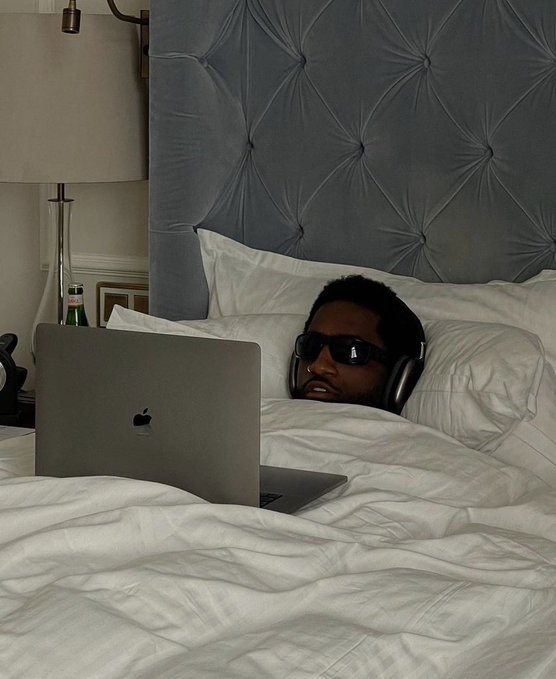 A person wearing sunglasses and headphones lies in bed with white sheets against a tufted grey headboard, working on a silver Apple MacBook laptop.