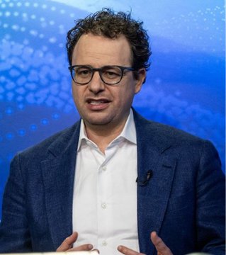 A man with curly hair and glasses wearing a dark blazer over a white shirt, speaking with hand gestures against a blue background.