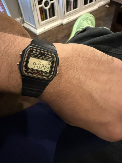 A close-up of a wrist shows a black Casio F-91W digital watch displaying “TU 20” and “9:02 23,” with labels including “CASIO,” “ALARM CHRONOGRAPH,” “WATER,” “WR,” and “RESIST.”
