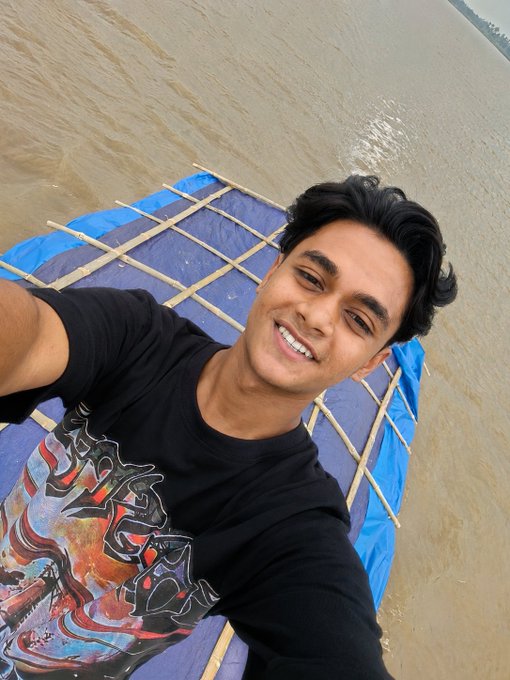 A young man in a black t-shirt smiles for a selfie taken from an elevated angle on a structure over sandy beach waters.