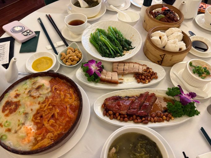 A table full of traditional Hong Kong dim sum and dishes, including steamed dumplings, roasted meats, sautéed greens, and congee, shared among friends during a long lunch. A pink smartphone case with "03" and a numbered receipt are visible on the table, matching the post about enjoying authentic Cantonese cuisine while discussing industry topics and US stock options trading.