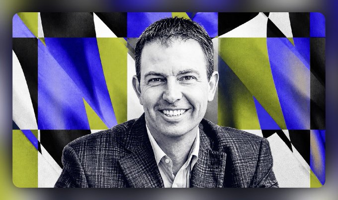 A black and white photo of a smiling man in a checkered blazer set against a vibrant abstract background of blue, yellow, black, and white geometric shapes.
