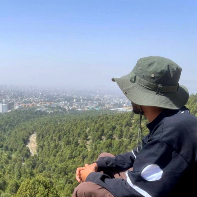 A man wearing a green bucket hat sits on a high vantage point, gazing out over a vast forested landscape and a distant cityscape.