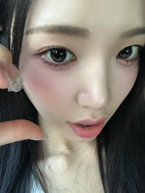 An extreme close-up selfie of a young woman with dark hair, soft pink blush, eyeliner, and glossy lips, with her finger resting near her cheek.
