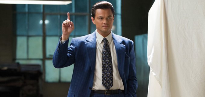 A man in a blue pinstripe suit and patterned tie stands in a room, pointing his index finger upward in a defiant gesture.