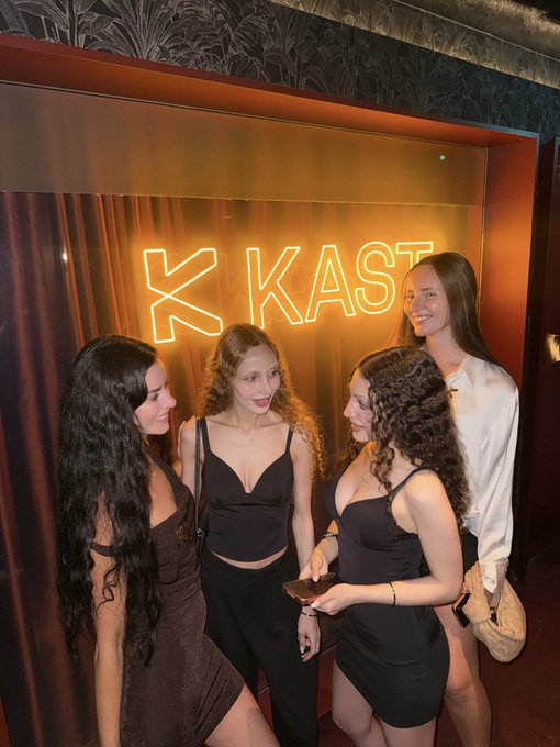 Four women posing together in front of a neon sign that reads 'K KAST' in an orange-lit venue.