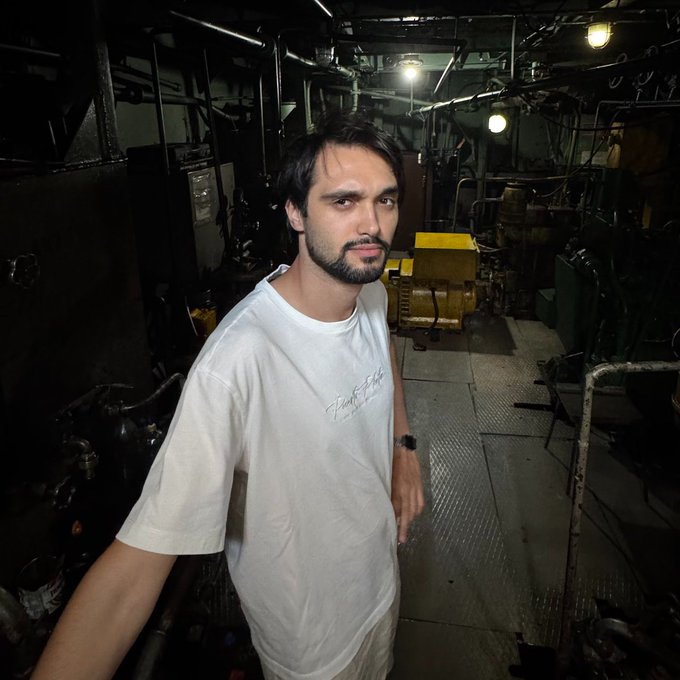 A selfie portrait of a bearded man in a white t-shirt standing in what appears to be the engine room of a ship.