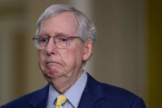 Senator Mitch McConnell, wearing glasses, a navy suit, and yellow tie, appears with a serious expression during a press event related to his vote in favor of advancing the "Crypto Clarity Act."