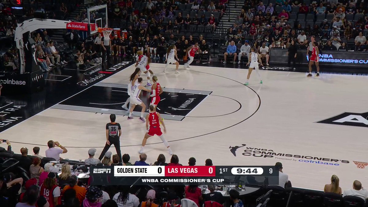 The image shows a basketball game with players on the court, including one player wearing a red shirt who has just made a shot. The crowd appears to be watching intently as the game unfolds. In the background, there are advertisements for various companies such as #varsitycombs, #wnascombs, and #coinbase.